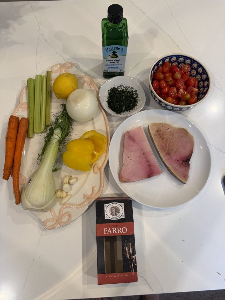 Marble counter with carrots, celery, fennel, lemon, onion, a yellow pepper, garlic, dill, grape tomatoes, olive oil, two fish steaks and a box of farro